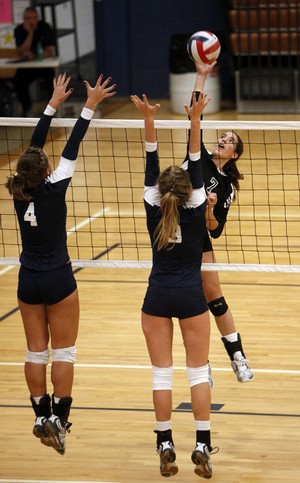 high school girl attempting to spike with 2 defenders on the opposite side of the net all wearing black official standard equipment volley ball