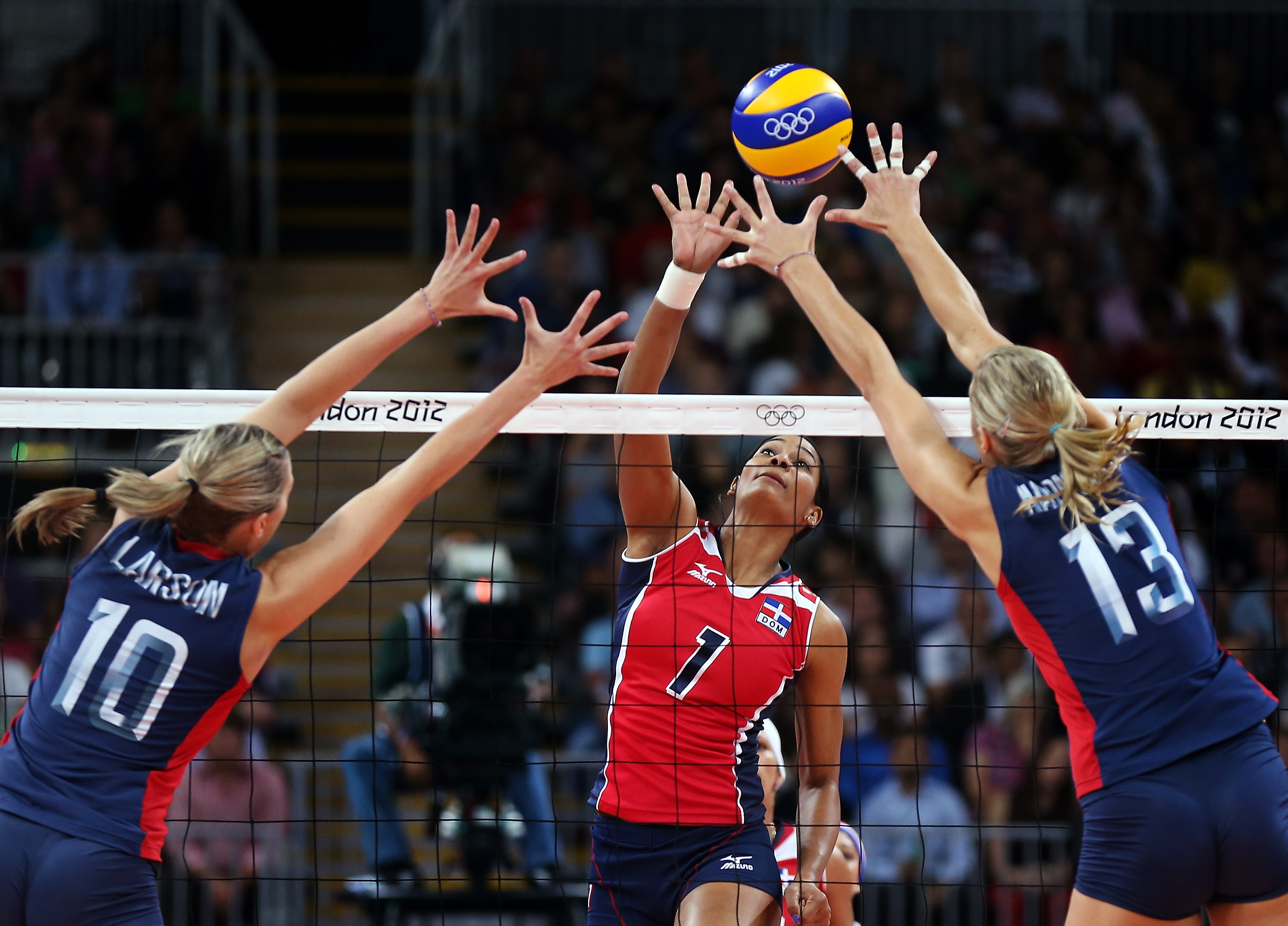 woman in red spiking the ball while 2 women in blue try to block the ball basic equipment for volleyball ball is a regulated ball