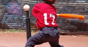 little kid swinging a bat at the tee ball ball on the tee ball stand red shirt helmet basic equipment for tee ball