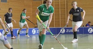 girl in green uniform running up floorball court with a player in black following standard equipment for floorball