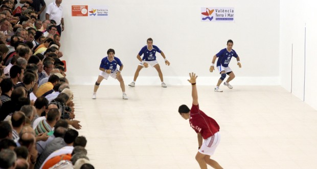 traditional valencian pilota game with 3 men in blue while a man in red is about to sweep down to hit the ball standard equipment for valencian pilota game