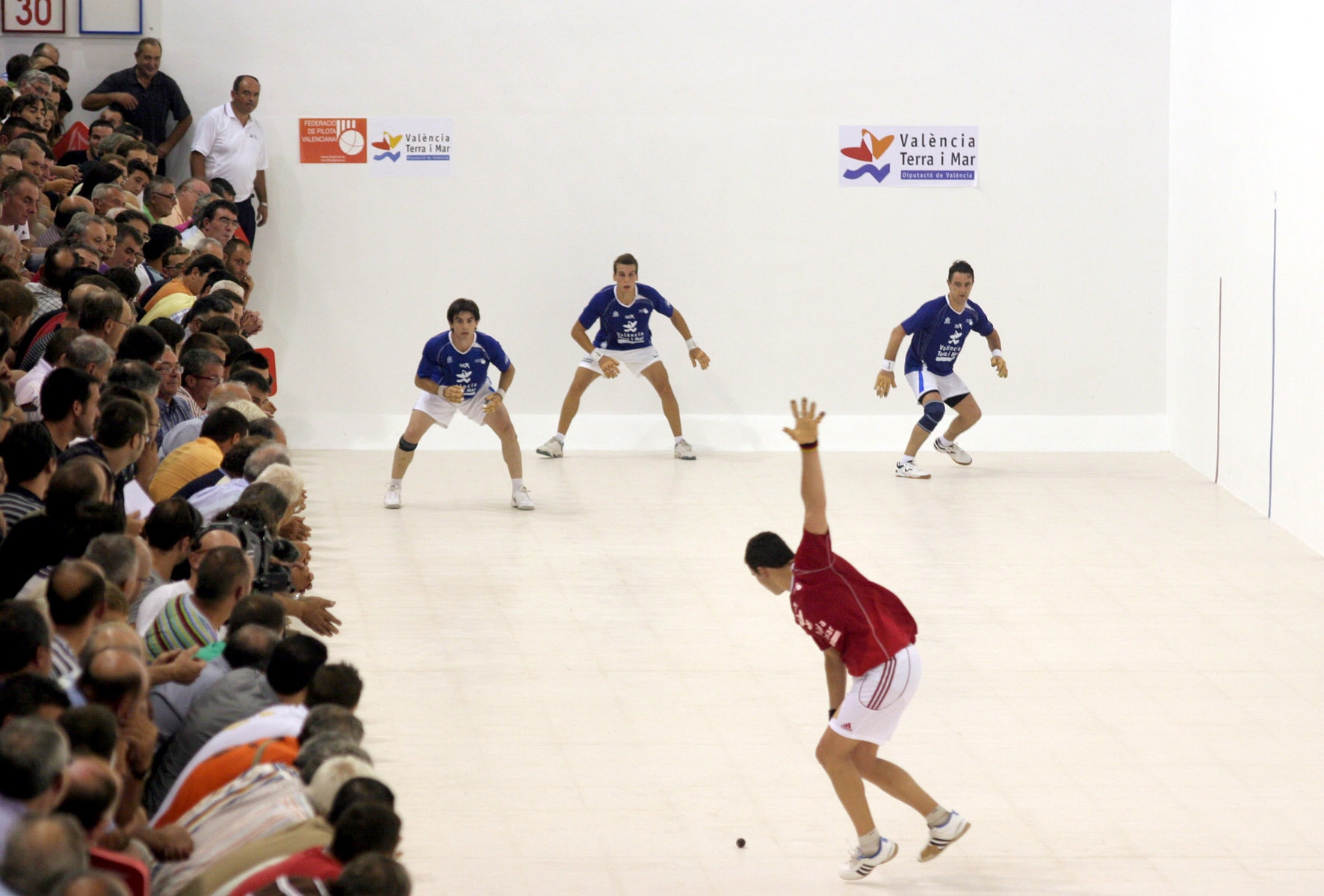 traditional valencian pilota game with 3 men in blue while a man in red is about to sweep down to hit the ball standard equipment for valencian pilota game