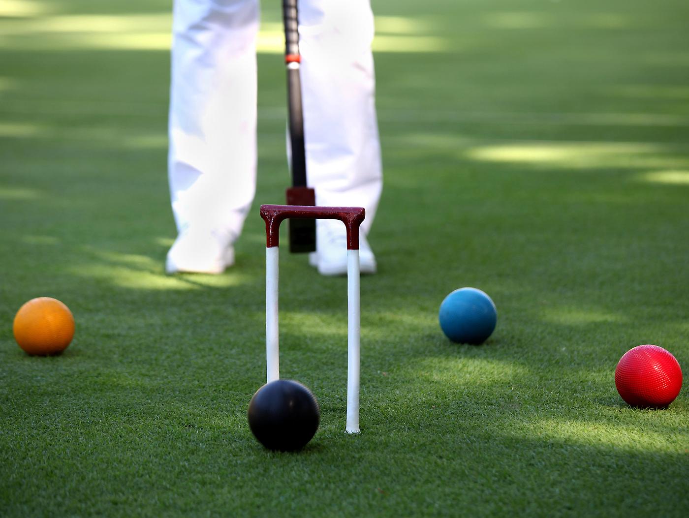 person in white pants standing behind a croquet game with orange/yellow ball black ball blue ball and red ball standard equipment for croquet